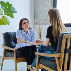 Terapeuta w okularach słucha pacjentki, która opowiada o swoich trudnościach. W tle duża roślina.