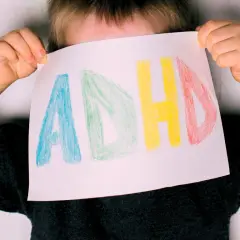 Dziecko z ADHD trzyma kartkę z napisem "ADHD" narysowanym kolorowymi kredkami.