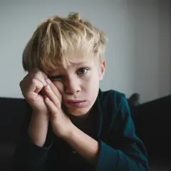 Chłopiec z blond włosami, zamyślony, opiera głowę na dłoniach. Jego wyraz twarzy może sugerować niektóre objawy aspergera, jak trudności w wyrażaniu emocji.