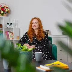 Kobieta z rudymi włosami w czarnej sukience w kropki siedzi w zielonym fotelu. Na stoliku leżą książki, a w tle widać regał z dekoracjami. To idealne miejsce na rozmowy z conners 3.