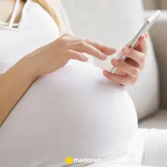Kobieta w ciąży, w białej bluzce i beżowym kardiganie, przegląda coś na telefonie. To może być jej l4 ciążowe.