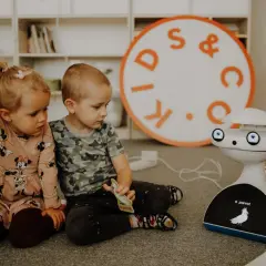 Dwoje dzieci, chłopiec i dziewczynka, siedzą na dywanie, bawiąc się z robotem. To piękny obraz dzieciństwa, pełen ciekawości i nauki.
