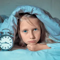 Smutna dziewczynka z niebieskimi oczami leży pod kołdrą obok budzika. Zmaga się z zaburzeniami snu.