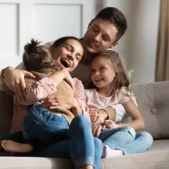 Szczęśliwa rodzina na kanapie. Mama śmieje się, tuląc córkę. Tata obejmuje drugą córkę. To piękny obraz rodziny, gdzie każdy czuje się kochany, nawet jeśli ktoś jest pasierbem, kto to nie ma znaczenia w tej miłości.