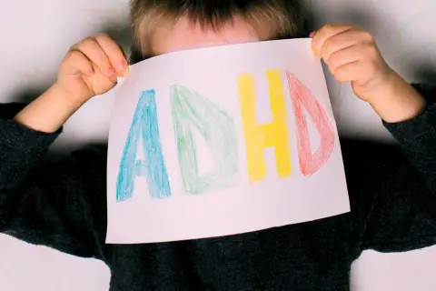 Dziecko z ADHD trzyma kartkę z napisem "ADHD" narysowanym kolorowymi kredkami.
