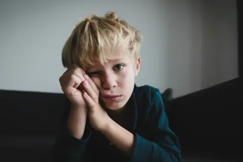 Chłopiec z blond włosami, zamyślony, opiera głowę na dłoniach. Jego wyraz twarzy może sugerować niektóre objawy aspergera, jak trudności w wyrażaniu emocji.