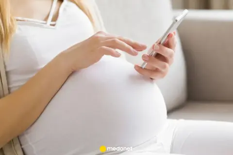 Kobieta w ciąży, w białej bluzce i beżowym kardiganie, przegląda coś na telefonie. To może być jej l4 ciążowe.