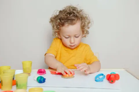 Chłopiec z kręconymi włosami bawi się plasteliną, tworząc kształty. Jego skupienie może być oznaką cech związanych z aspergerem u dzieci.