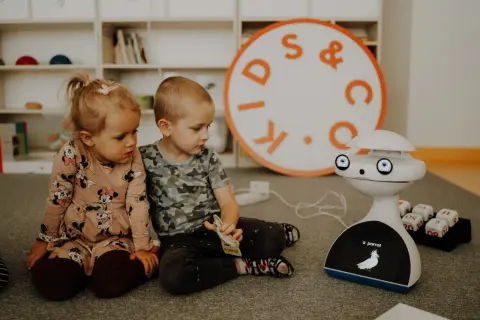 Dwoje dzieci, chłopiec i dziewczynka, siedzą na dywanie, bawiąc się z robotem. To piękny obraz dzieciństwa, pełen ciekawości i nauki.