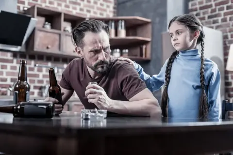 Smutna dziewczynka kładzie rękę na ramieniu ojca, który pije alkohol. To obraz trudnej sytuacji rodzinnej, która może wymagać terapii DDA.