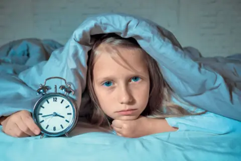 Smutna dziewczynka z niebieskimi oczami leży pod kołdrą obok budzika. Zmaga się z zaburzeniami snu.