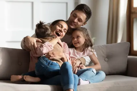 Szczęśliwa rodzina na kanapie. Mama śmieje się, tuląc córkę. Tata obejmuje drugą córkę. To piękny obraz rodziny, gdzie każdy czuje się kochany, nawet jeśli ktoś jest pasierbem, kto to nie ma znaczenia w tej miłości.