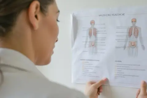 Lekarka analizuje schemat układu kostnego, zastanawiając się, czy do neurologa potrzebne jest skierowanie.