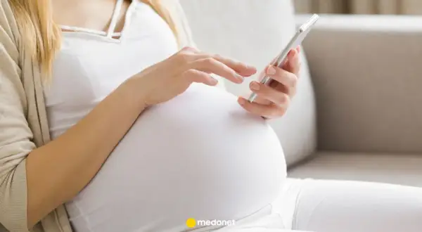 Kobieta w ciąży, w białej bluzce i beżowym kardiganie, przegląda coś na telefonie. To może być jej l4 ciążowe.