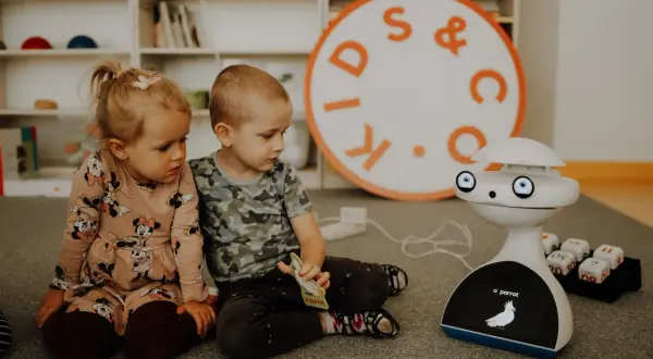 Dwoje dzieci, chłopiec i dziewczynka, siedzą na dywanie, bawiąc się z robotem. To piękny obraz dzieciństwa, pełen ciekawości i nauki.