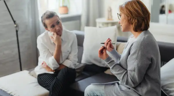 Psycholog i psychiatra rozmawiają z pacjentką w gabinecie. Kobieta w białej koszuli słucha uważnie, opierając podbródek na dłoni.