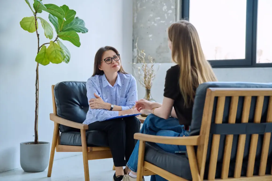 Terapeuta w okularach słucha pacjentki, która opowiada o swoich trudnościach. W tle duża roślina.