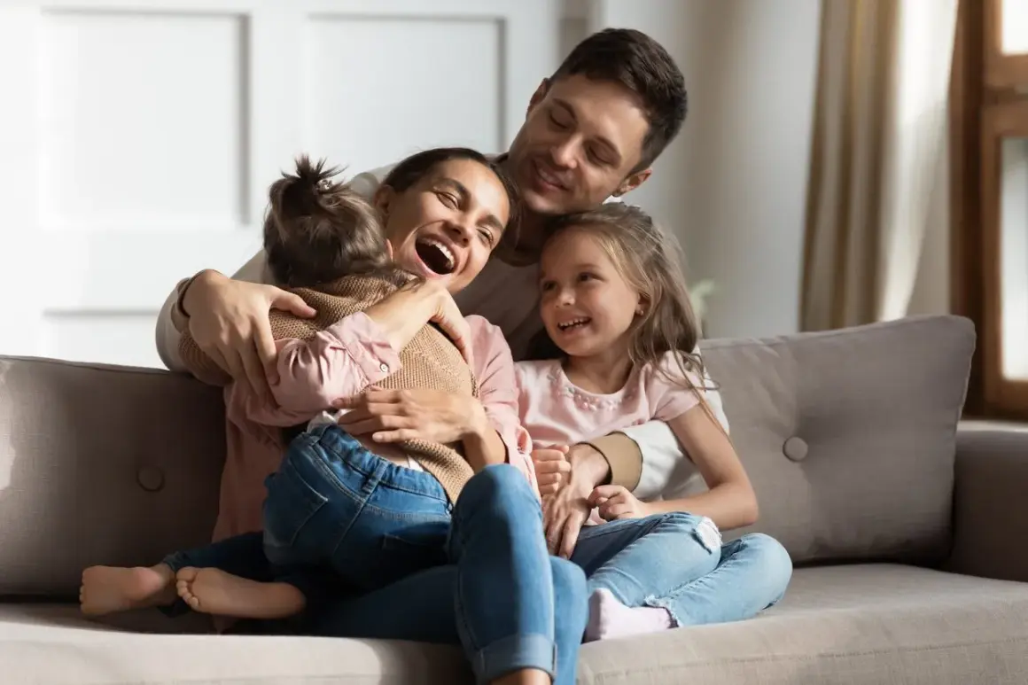 Szczęśliwa rodzina na kanapie. Mama śmieje się, tuląc córkę. Tata obejmuje drugą córkę. To piękny obraz rodziny, gdzie każdy czuje się kochany, nawet jeśli ktoś jest pasierbem, kto to nie ma znaczenia w tej miłości.