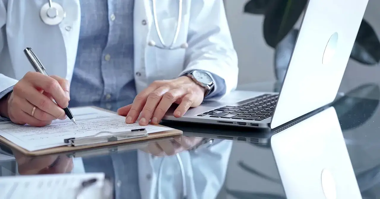 Lekarz w białym kitlu pisze na dokumentach, obok laptop. Może to być dokumentacja medyczna, np. l4 od psychiatry.