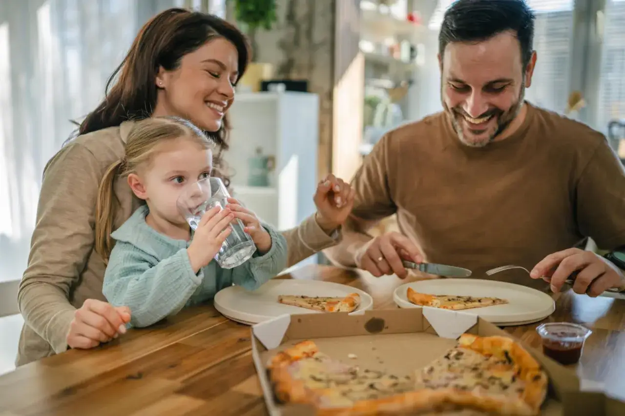 Szczęśliwa rodzina przy stole, córka pije wodę, a rodzic kroi pizzę.