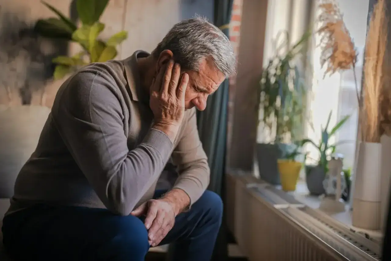 Starszy mężczyzna siedzi ze spuszczoną głową, opierając dłoń na twarzy. Jego postawa sugeruje głębokie stany depresyjne i przygnębienie.