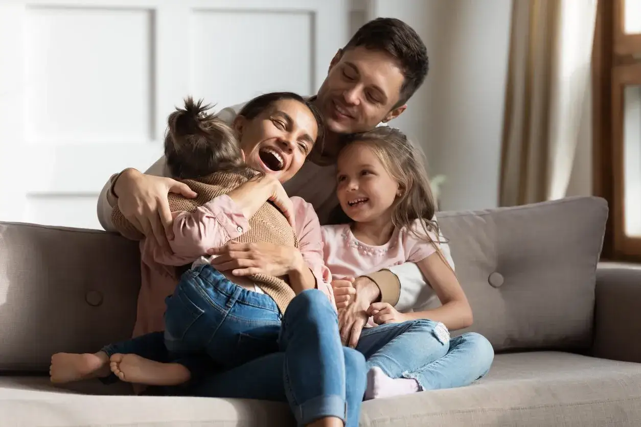 Szczęśliwa rodzina na kanapie. Mama śmieje się, tuląc córkę. Tata obejmuje drugą córkę. Wszyscy są uśmiechnięci, tworząc ciepłą atmosferę.