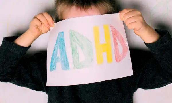 Dziecko z ADHD trzyma kartkę z napisem "ADHD" narysowanym kolorowymi kredkami.