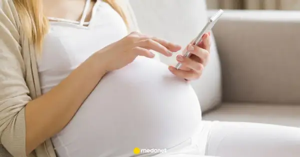 Kobieta w ciąży, w białej bluzce i beżowym kardiganie, przegląda coś na telefonie. To może być jej l4 ciążowe.